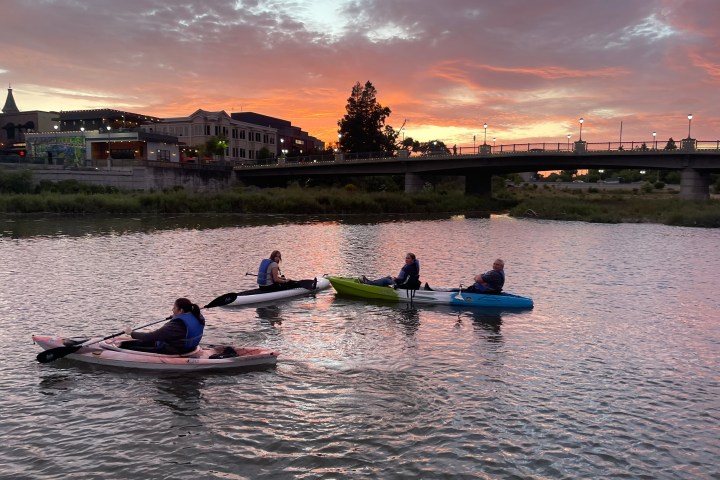 Four kayakers on a river at sunset with a bridge and buildings in the background.